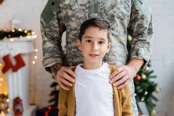 Hombre en uniforme militar abrazando hombros de hijo mirando a la cámara en casa - foto de stock