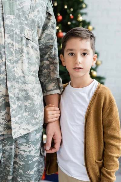 Kid looking at camera while holding hand of dad in camouflage at home — Stock Photo