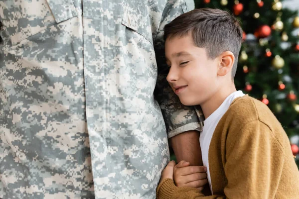 Niño sonriente con los ojos cerrados abrazando la mano del padre en uniforme militar - foto de stock