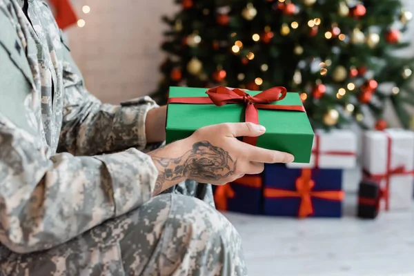 Vista cortada de soldado tatuado segurando caixa de presente verde com fita vermelha perto de árvore de natal borrada — Fotografia de Stock