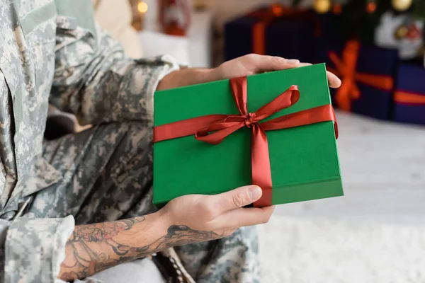 Vue partielle de l'homme en camouflage tenant boîte cadeau verte avec ruban rouge à la maison — Photo de stock