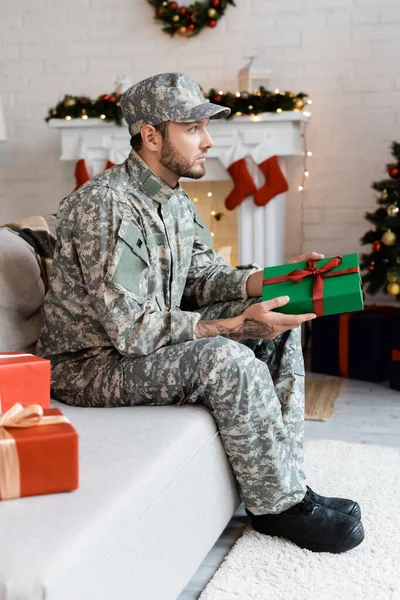 Side view of thoughtful man in military uniform holding christmas present and looking away on couch at home — Stock Photo