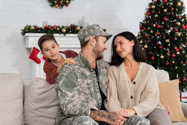 Menino alegre olhando para a câmera perto de mãe e pai em uniforme militar de mãos dadas no sofá perto da árvore de natal — Fotografia de Stock