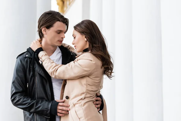 Bella giovane donna abbracciare fidanzato in giacca nera elegante vicino a colonne bianche — Foto stock