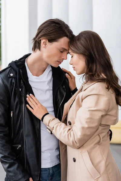 Young woman in beige trench coat hugging boyfriend in stylish jacket near white columns — Stock Photo