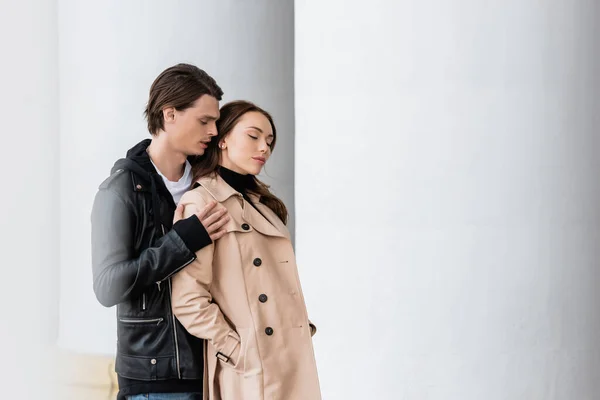 Stylischer Mann in Jacke umarmt Schultern einer sinnlichen Freundin, die mit Händen in Trenchcoat-Taschen in der Nähe von Säulen steht — Stockfoto