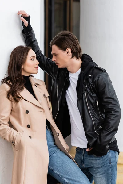 Homme élégant en veste noire flirtant avec jolie femme en trench beige debout avec la main dans la poche près de la colonne blanche — Photo de stock