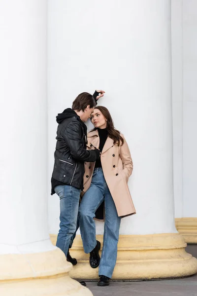 Pleine longueur de jeune homme en veste élégante flirtant avec jolie femme en trench-coat beige debout avec la main dans la poche — Photo de stock