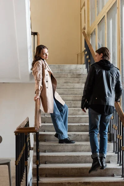 Full length of young man in stylish outfit looking at pretty girlfriend in beige trench coat standing on stairs — Stock Photo
