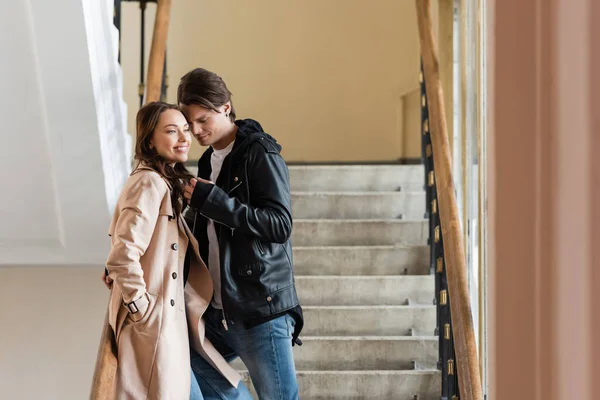 Junger Mann in stylischer Jacke flirtet mit fröhlicher Freundin im beigefarbenen Trenchcoat stehend mit der Hand in der Tasche — Stockfoto