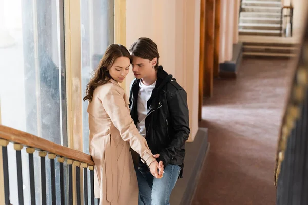 Joven en chaqueta negra coqueteando con su novia en abrigo otoñal mientras toma las manos en las escaleras - foto de stock