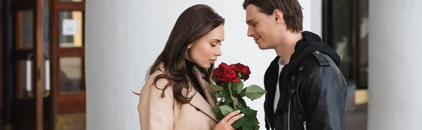 Side view of happy man smiling near pretty girlfriend smelling roses near white column, banner — Stock Photo