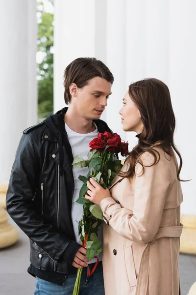 Pretty young woman holding red roses and looking at stylish boyfriend in jacket — Stock Photo