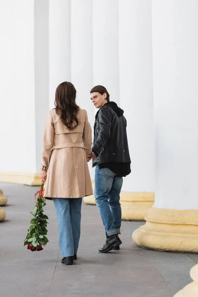 Piena lunghezza di felice giovane uomo che tiene la mano della ragazza in trench in piedi con rose rosse — Foto stock