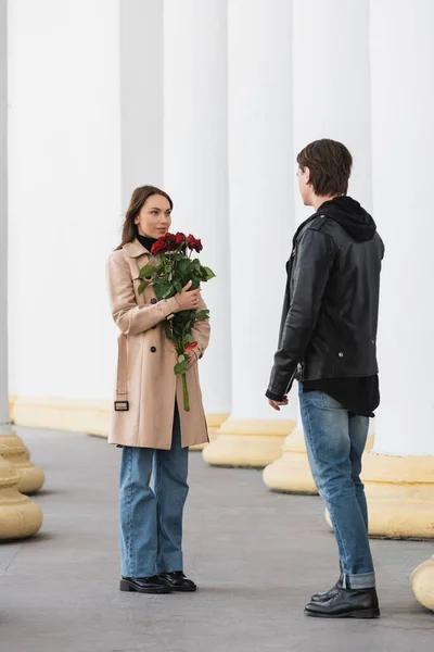 Longitud completa de la mujer joven bonita sosteniendo rosas rojas y mirando novio con estilo en chaqueta - foto de stock