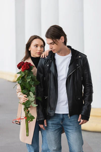 Bonita mujer joven sosteniendo rosas rojas y abrazando hombro de novio con estilo en la chaqueta - foto de stock