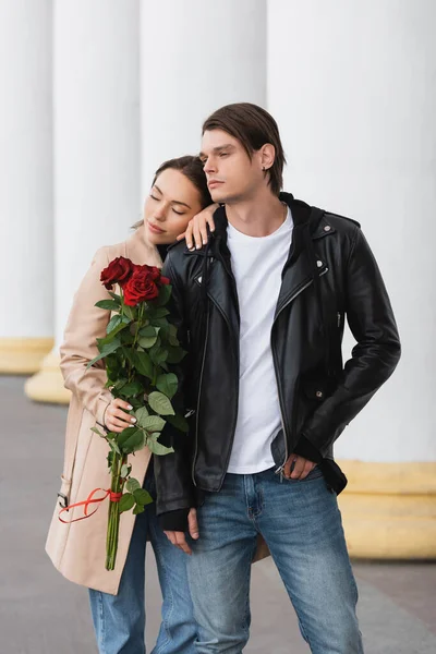 Bonita mulher segurando rosas vermelhas e inclinando-se no ombro do namorado elegante na jaqueta — Fotografia de Stock