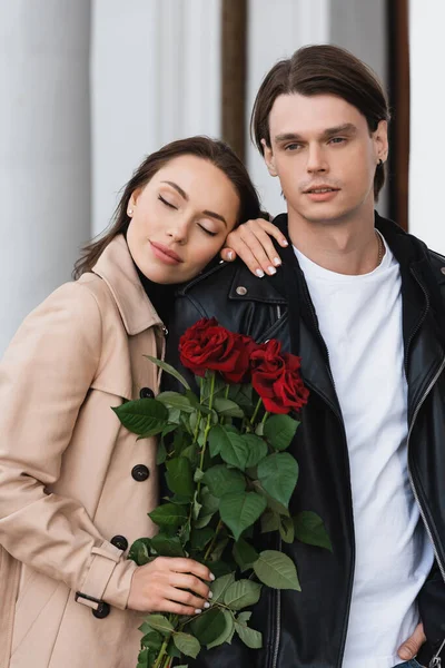 Pretty woman in trench coat holding red roses and leaning on shoulder of boyfriend in stylish jacket — Stock Photo