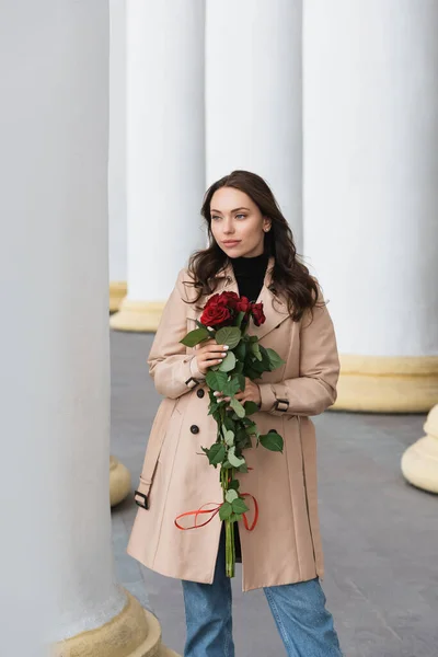 Jolie jeune femme en trench beige tenant des roses rouges et détournant les yeux — Photo de stock