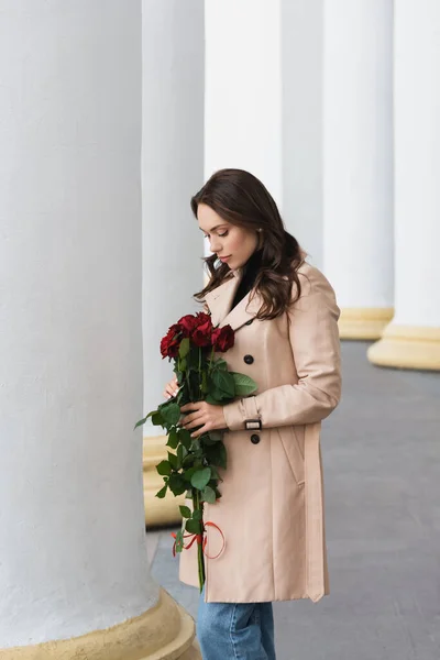 Bella giovane donna in trench beige guardando fioritura rose rosse — Foto stock