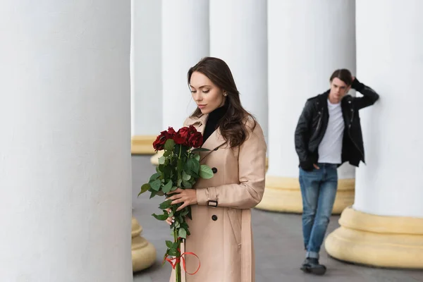 Bonita mujer joven en gabardina beige mirando rosas rojas cerca de novio sobre fondo borroso - foto de stock