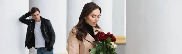 Bella giovane donna in trench beige guardando rose rosse vicino fidanzato su sfondo sfocato, banner — Foto stock