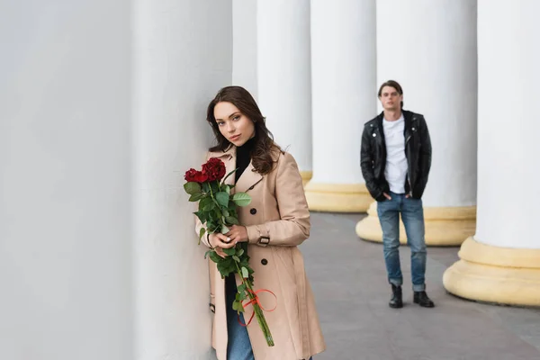 Hübsche Frau im beigen Trenchcoat mit roten Rosen in der Nähe ihres Freundes auf verschwommenem Hintergrund — Stockfoto