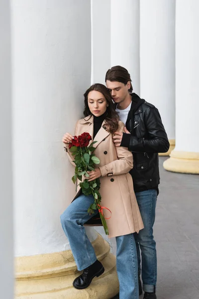 Повнометражний стильний молодий чоловік обіймає красиву жінку в бежевому траншеї пальто тримає червоні троянди — стокове фото