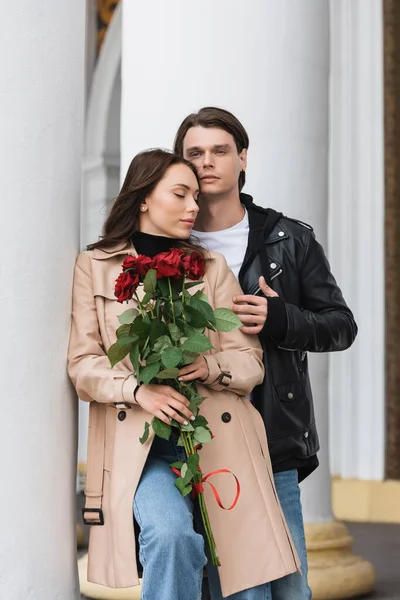 Elegante hombre abrazando bonita novia en elegante gabardina sosteniendo rosas rojas - foto de stock