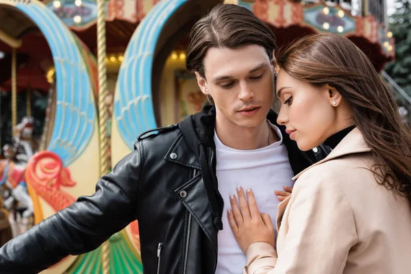 Young woman in beige trench coat hugging man near carousel in amusement park — Stock Photo