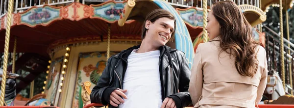 Homem elegante na jaqueta sorrindo perto da namorada feliz no casaco de trincheira e carrossel no parque de diversões, banner — Fotografia de Stock