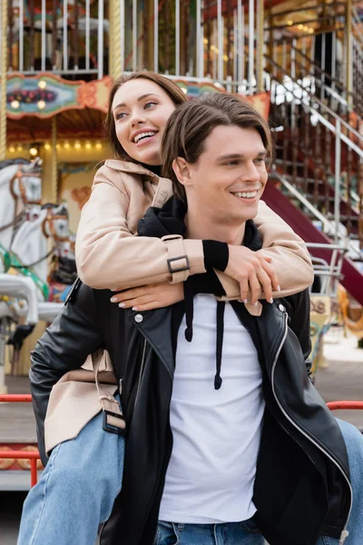 Homme gai dans la veste noire piggybackking petite amie heureuse dans le parc d'attractions — Photo de stock