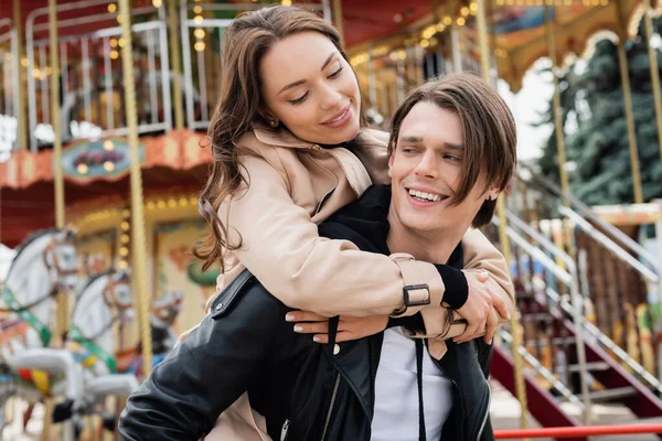 Mujer feliz en gabardina abrazando novio alegre en parque de atracciones - foto de stock