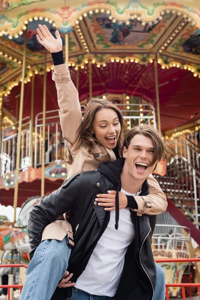 Excited man in black jacket piggybacking happy girlfriend in amusement park — Stock Photo