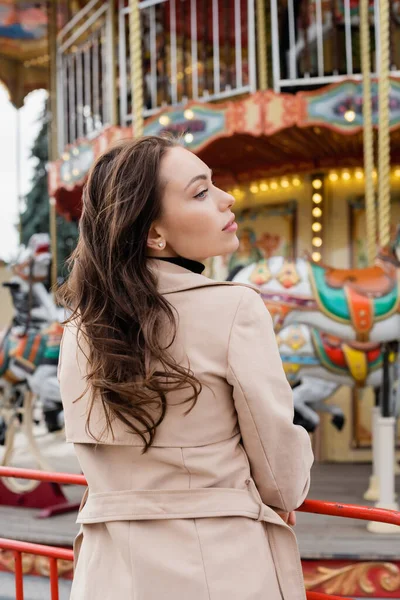 Curly woman in beige trench coat and jeans standing near carousel in amusement park — Stock Photo