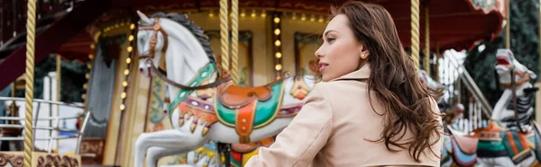 Side view of curly young woman in beige trench coat standing near carousel in amusement park, banner — Stock Photo