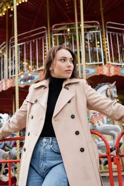 Brunette young woman in beige trench coat and jeans standing near carousel in amusement park — Stock Photo