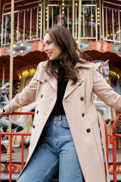 Cheerful young woman in beige trench coat and jeans standing near carousel in amusement park — Stock Photo