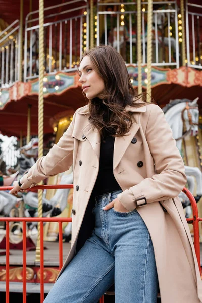Young woman in beige trench coat and jeans standing with hand in pocket near carousel in amusement park — Stock Photo