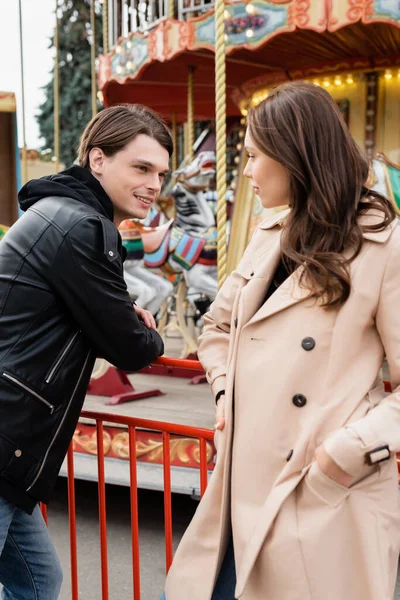 Homem alegre olhando para a namorada em casaco de trincheira de pé com as mãos nos bolsos no parque de diversões — Fotografia de Stock