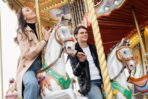 Happy young couple holding hands and riding carousel horses in amusement park — Stock Photo