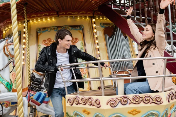 Hombre alegre en traje de moda mirando novia feliz con las manos en alto carrusel en el parque de atracciones - foto de stock