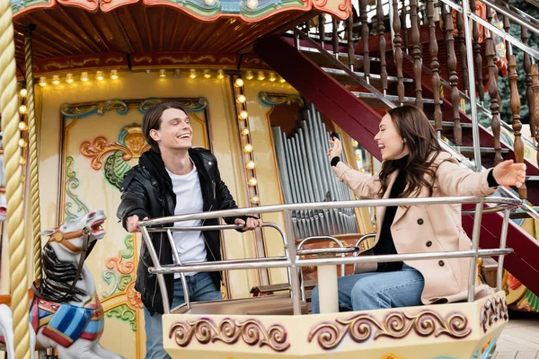 Alegre hombre en elegante traje mirando feliz novia riendo en carrusel en parque de atracciones - foto de stock