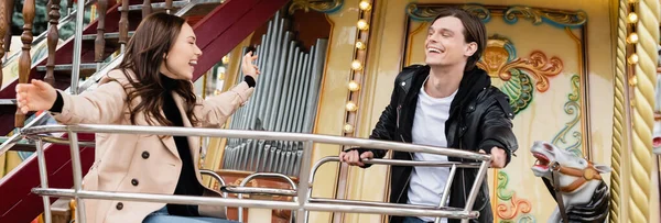 Hombre alegre en traje elegante mirando novia feliz riéndose en carrusel en parque de atracciones, bandera - foto de stock