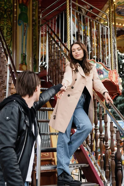 Young man in stylish jacket holding hand of pretty girlfriend on carousel in amusement park — Stock Photo