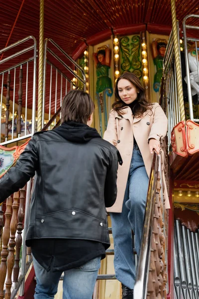 Young man in stylish jacket holding hand of girlfriend on carousel in amusement park — Stock Photo