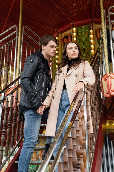 Vista de ángulo bajo de pareja elegante cogida de la mano mientras está de pie en el carrusel en el parque de atracciones - foto de stock