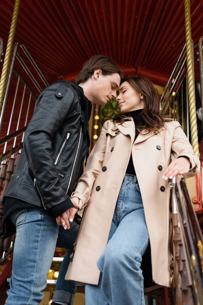 Vista de ángulo bajo de pareja sensual cogida de la mano mientras está de pie en el carrusel en el parque de atracciones - foto de stock