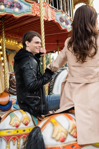 Happy young man holding hands with girlfriend and riding carousel horses in amusement park — Stock Photo