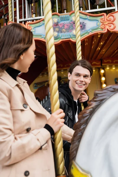 Heureux jeune homme regardant petite amie équitation carrousel cheval dans le parc d'attractions — Photo de stock